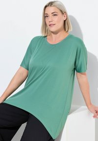Haut vert à manches courtes en tissu lisse et léger, avec une coupe décontractée, doté d'un col rond et de fentes sur les côtés. Porté par-dessus un pantalon noir.