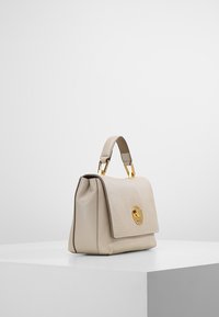 Beige nahkalaukku ylhäällä olevalla kahvalla ja kultasävyisellä soljella, esillä valkoisella jalustalla yksinkertaista taustaa vasten.