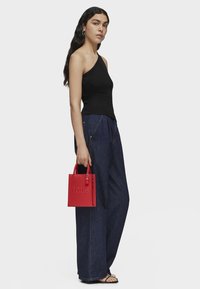 Top negro de un hombro, pantalones de mezclilla azules sueltos, bolso de cuero rojo con el logo en relieve "TOUS" y sandalias negras con tiras.