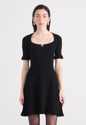Une jeune femme se tient avec assurance, vêtue d'une robe noire ajustée à manches courtes avec un détail en forme de cœur au niveau de la clé, et des boucles d'oreilles pendantes.