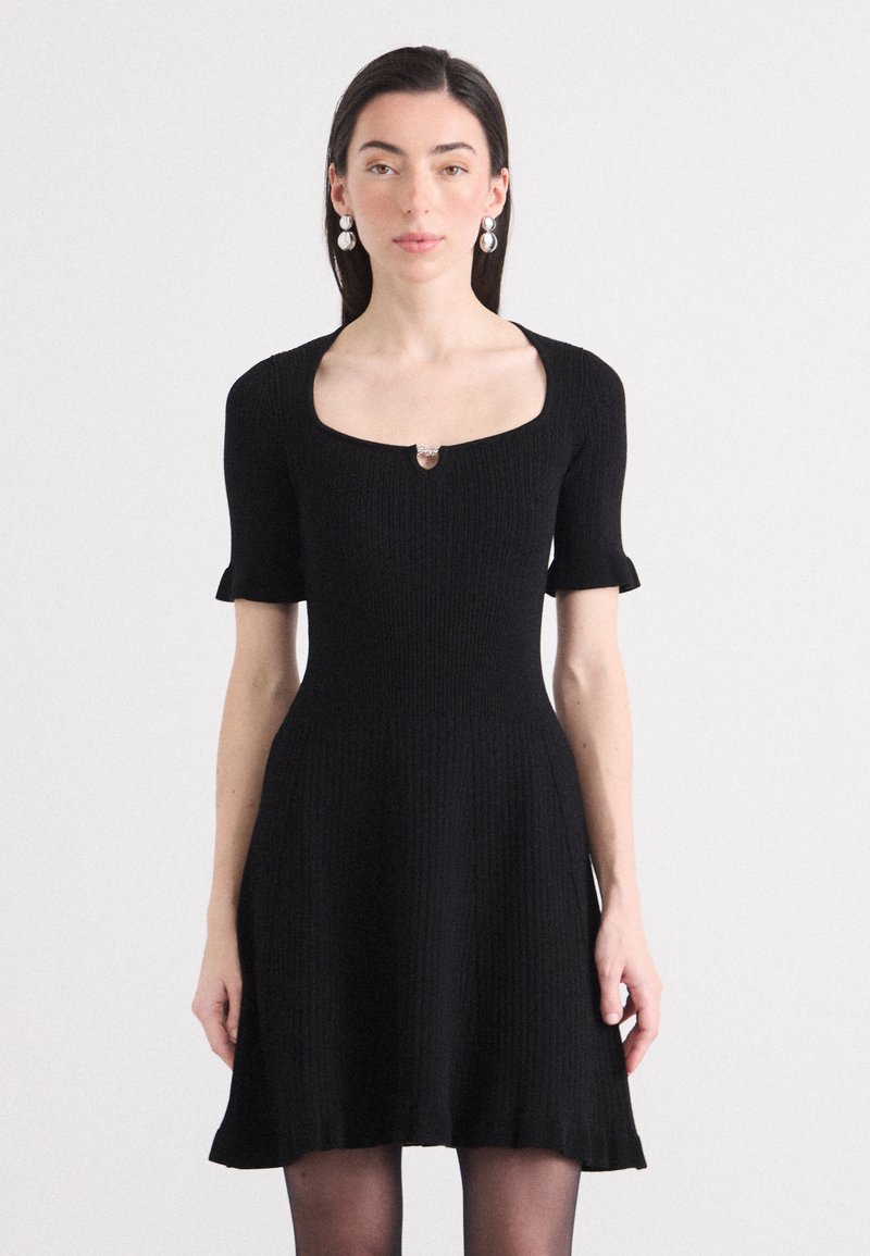 A young woman stands confidently, wearing a fitted black dress with short sleeves and a heart-shaped keyhole detail, with dangling earrings.