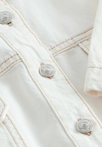 Tessuto di denim bianco con cuciture in filo di rame, dotato di bottoni metallici argentati con testo in rilievo. Ingrandimento del colletto e della patta dei bottoni.