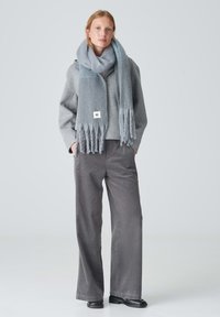 Sciarpa di lana grigia con bordi frangiati appoggiata intorno al collo di un modello, abbinata a una giacca oversize grigia e pantaloni larghi grigi.