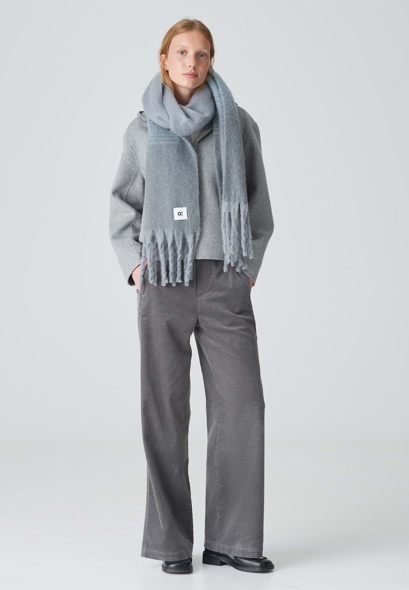 Sciarpa di lana grigia con bordi frangiati appoggiata intorno al collo di un modello, abbinata a una giacca oversize grigia e pantaloni larghi grigi.