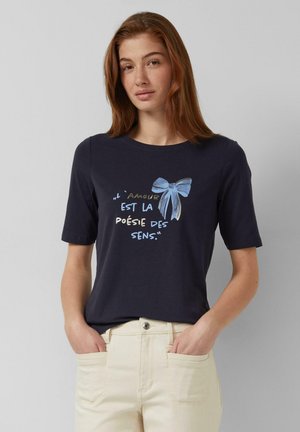 Junge Frau mit rotem Haar trägt ein marineblaues Shirt mit französischem Text und einer blauen Schleifen-Grafik, kombiniert mit hellbeigen Hosen, die Hände in den Taschen.