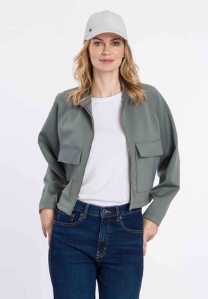 Giacca verde chiaro con zip e due tasche frontali, indossata sopra una maglietta bianca e abbinata a jeans blu scuro, completata da un cappellino grigio chiaro.