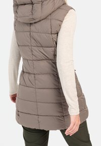 Gilet imbottito e con cappuccio in marrone spento con trapuntatura orizzontale. Presenta un collo alto e zip nelle tasche laterali. Tessuto morbido.