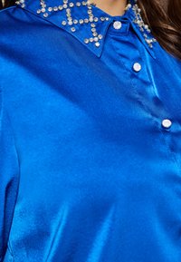 Satin blauwe shirt met een kraag versierd met heldere strassstenen en parelachtige accenten, met een glanzende, gladde textuur en knoopsluitingen.