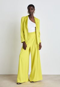 Gele op maat gemaakte blazer en wijde broek, gecombineerd met een witte top. Glad materiaal, strakke vormgeving en subtiele plooien op de broek.