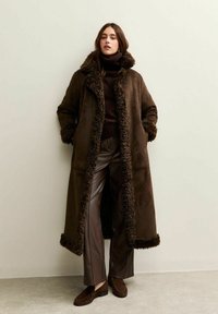 Vrouw in een lange, donkerbruine shearling jas, bruine coltrui, leren broek en bruine loafers, staande tegen een effen muur.