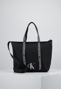 Czarna torba tote Calvin Klein z logo z przodu, dwoma markowymi uchwytami i długim regulowanym paskiem na ramię na białej powierzchni wystawowej.