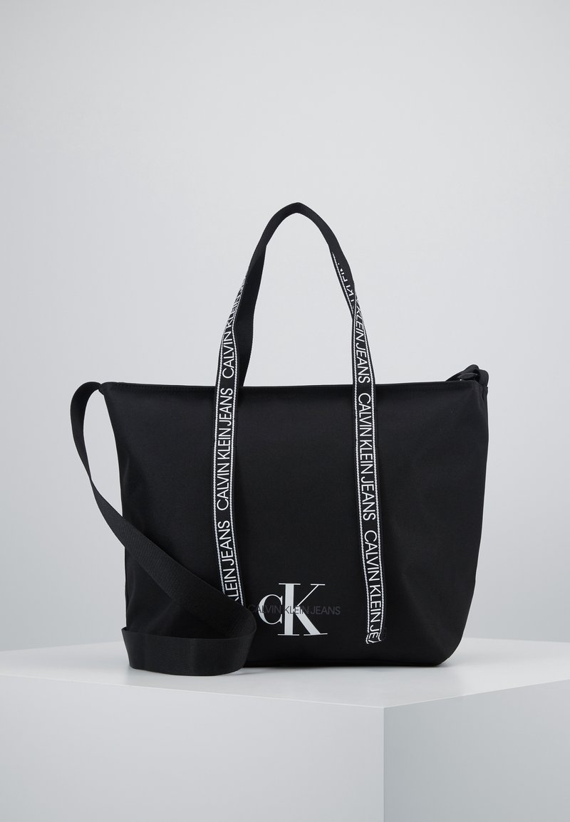 Czarna torba tote Calvin Klein z logo z przodu, dwoma markowymi uchwytami i długim regulowanym paskiem na ramię na białej powierzchni wystawowej.