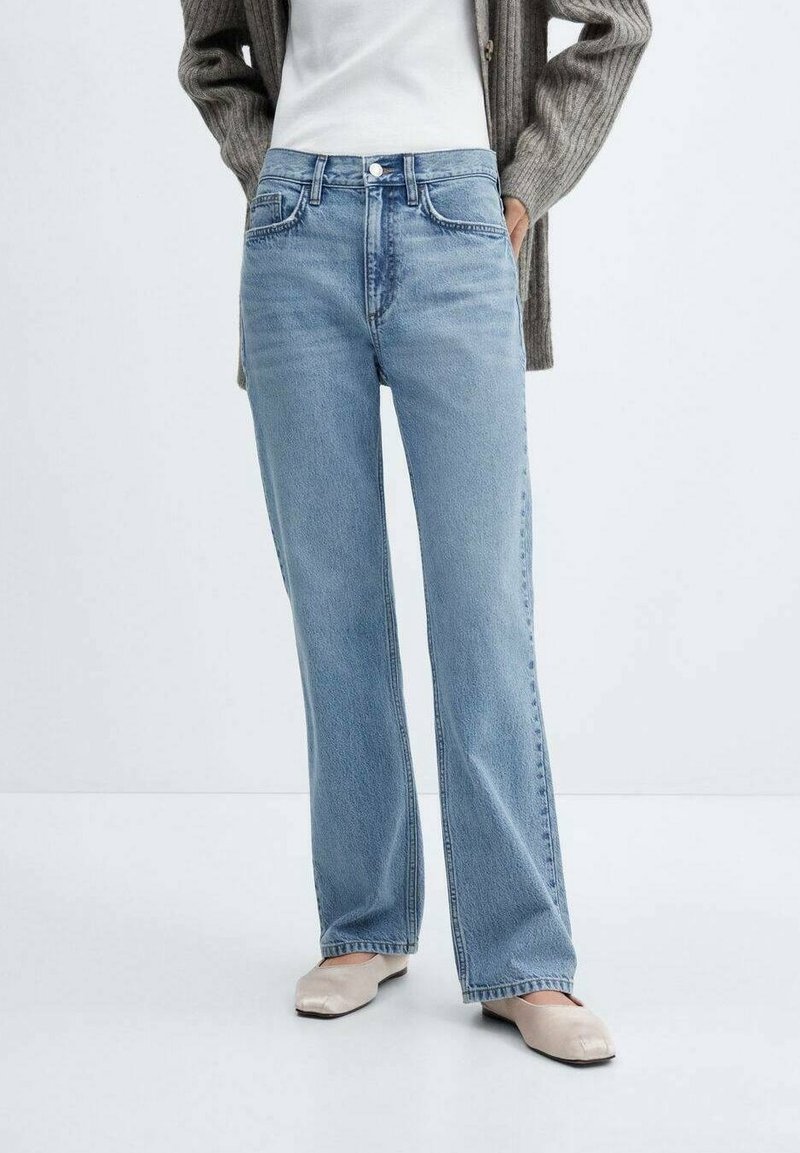 Personne portant un jean droit bleu clair, une chemise blanche, un cardigan gris et des chaussures plates beiges, debout devant un fond uni.