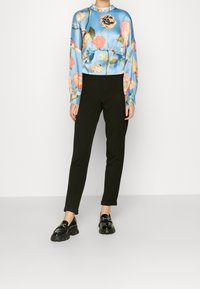 Person som bär svarta slim fit-byxor, svarta chunky loafers och en blå långärmad blus med rosa och gula blommönster.