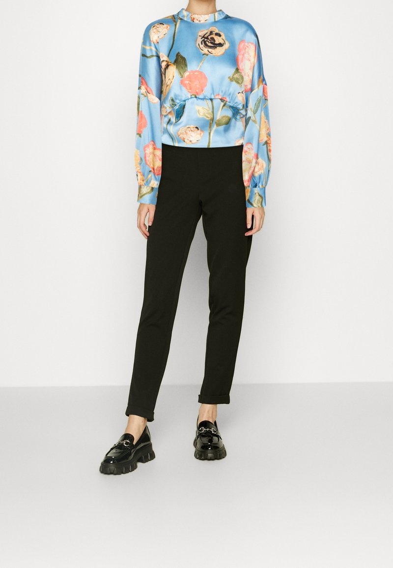 Person som bär svarta slim fit-byxor, svarta chunky loafers och en blå långärmad blus med rosa och gula blommönster.