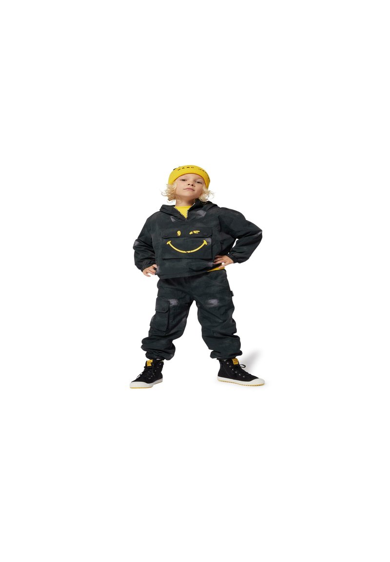 Zwarte camouflage jumpsuit met een gele glimlach, gecombineerd met een gele beanie. De outfit heeft cargozakken en zwarte schoenen.