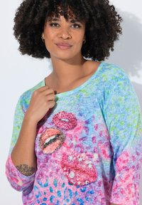 Frau mit lockigem Haar, die ein farbenfrohes blaues und pinkes Hemd mit Pailletten verzierten Lippenmotiven trägt und ein florales Tattoo an ihrem Unterarm zeigt.