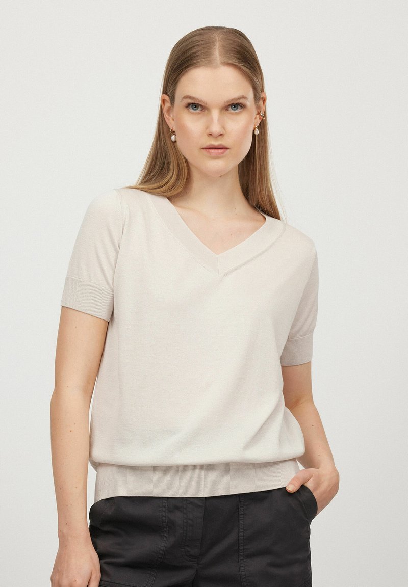 Lys beige kortærmet V-hals trøje med en glat tekstur og ribbede manchetter, parret med mørke bukser. Enkelt design, ingen mønstre eller accenter.