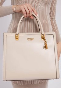 Sac à main en cuir beige avec une forme structurée, accents en métal doré et un porte-clé. Les poignées sont lisses et le sac arbore un logo à l'avant.