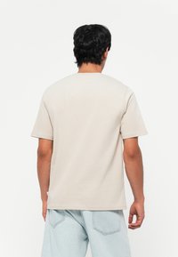 Beige cotton t-shirt with short sleeves, crew neck, and a relaxed fit. Features stitching details and a tag at the lower left seam.