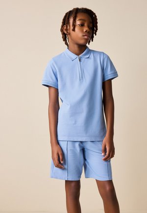 Ung person med korte dreadlocks iført en lyseblå kortærmet polo og matchende shorts, stående mod en ensfarvet beige baggrund.