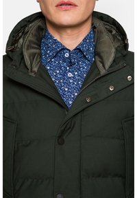 Ciemnozielona kurtka ocieplana o fakturowanym wykończeniu, z kapturem, zapięciem na zamek błyskawiczny i zatrzaskami, nałożona na niebieską koszulę w kwiaty.