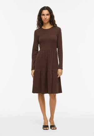 Femme debout vêtue d'une robe marron à manches longues et longueur genou, avec des sandales noires ouvertes au niveau des orteils, sur un fond blanc uni.