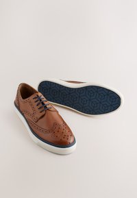 Braune Lederschuhe mit perforierten Details, blauen Schnürsenkeln und weißen Gummisohlen. Die Außensohle zeigt ein geometrisches Muster.