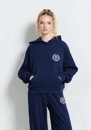 Blondynka z kucykiem, ubrana w granatową bluzę z kapturem i spodnie, oba z białym emblematem na piersi i udzie, stojąca na tle białej ściany.