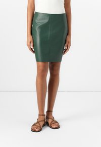 Zöld bőr ceruza szoknya slim fit szabással, sima textúrával és térd hosszúságú fazonnal. Barna lapos szandálokkal kombinálva, amelyek keresztbe tett pántokkal rendelkeznek.
