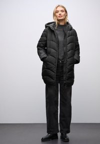 Cappotto puffo nero con cappuccio, caratterizzato da un motivo trapuntato, chiusura con zip e tasche laterali. Indossato sopra un maglione grigio e jeans scuri.