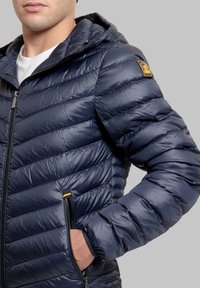 Giacca imbottita blu navy con cappuccio, caratterizzata da un design trapuntato, chiusura frontale con zip, due tasche laterali e un piccolo logo giallo sulla spalla.