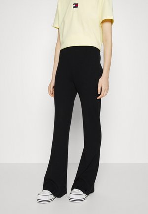 Persoon draagt een geel Tommy Jeans t-shirt, zwarte wijde hoge taille broek en witte platformsneakers, staand tegen een witte achtergrond.