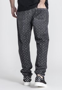 Pantalones de mezclilla negros con un patrón de logotipo repetido, cinco bolsillos y un corte relajado, combinados con zapatillas de deporte con un patrón en blanco y negro.