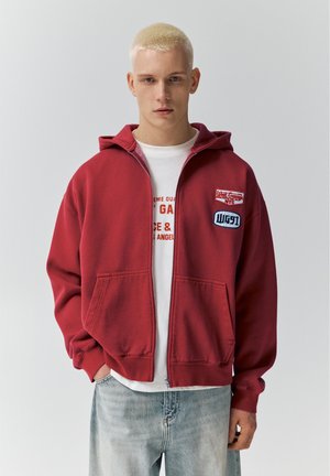 Młody mężczyzna z krótkimi blond włosami, ubrany w czerwoną bluzę na zamek z naszywkami, na białej koszulce z nadrukiem oraz w wyblakłe niebieskie dżinsy.