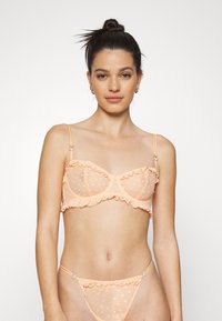 Conjunto de lingerie cor pêssego apresentando um bralette transparente com babados, padrão de bolinhas e alças ajustáveis. A tanga correspondente tem detalhes semelhantes.