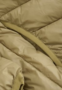 Olivgrön quiltad pufferjacka i ett tyg med en slät, glänsande yta. Har en kontrasterande matt tygkant längs sömmarna.
