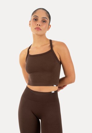 Kvinde stående med armene bag ryggen, iført en tætsiddende brun crop top og matchende højtaljede leggings mod en ensfarvet baggrund.