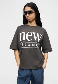 Γυναίκα που φοράει σκουρόχρωμο φαρδύ t-shirt "New Balance", μπλε υφασμάτινο παντελόνι και αθλητικά μαύρα γυαλιά ηλίου, στέκεται μπροστά σε απλό λευκό φόντο.