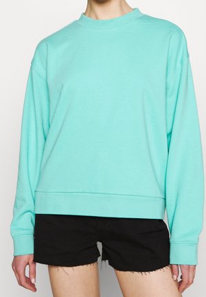 Personne portant un sweat-shirt vert menthe à manches longues et un short en jean noir effiloché, debout devant un fond clair uni.