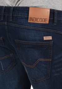 Tamno plave traperice s kožnom etiketom "INDICODE" na pojasu, kontrastnim narančastim šavovima i standardnim stražnjim džepovima.