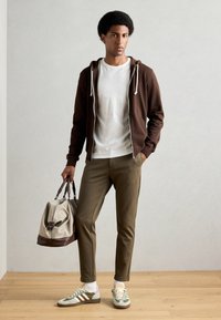 Sudadera marrón con cremallera, camiseta blanca, pantalones ajustados oliva, zapatillas verdes y grises con acentos rosas, llevando un bolso de lona bicolor.