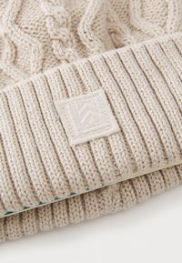 Strikket lue i lys beige med kabelmønster og ribbestrikket bånd. Inkluderer en liten, firkantet logo-patch med teksturdetaljer.