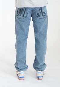 Jean en denim bleu clair avec des poches brodées uniques, portant les lettres 'CM' et 'A' en fil foncé. Coupe ample et texture subtile.