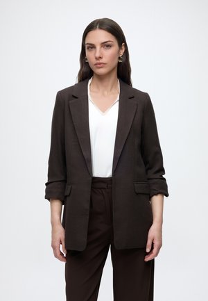 Femme aux longs cheveux foncés porte un blazer marron sur une blouse blanche et un pantalon marron, debout devant un fond blanc uni.