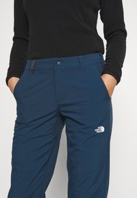 Blå utomhusbyxor gjorda av ett lätt material, med knäppning fram, sidofickor och en vit logotyp på nedre ben.