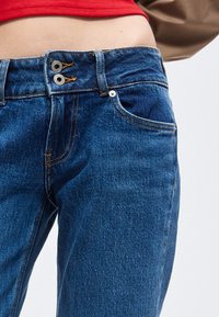 Suurelt võetud keskmise säärega sinised teksapüksid, millel on kahekordne nööpide kinnitamine ja nähtav punase särke ülaosa.