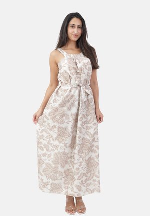 Femme portant une robe blanche sans manches et longue avec des motifs fleuris beiges, posant les mains tenant les côtés de la robe.