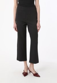 Pantaloni neri a gamba larga con vita elastica e texture liscia, abbinati a scarpe con tacco bordeaux a punta e con decorazioni metalliche.