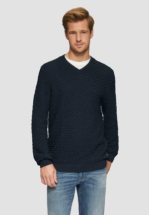 Marineblauer Strickpullover mit Diamantmuster, V-Ausschnitt, gerippten Bündchen und Saum, kombiniert mit hellblauen Jeans. Strukturiert gewebter Stoff.